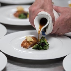 Close-up image of hands pouring a sauce onto a plate of salmon