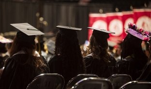 Four graduates are seen from behind, sitting in Harding Mazzotti Arena, at Commencement 2026