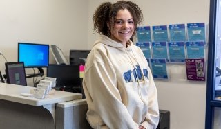 Sophia Goldsmith smiles at the camera in the Student Activities office. She has dark curly hair and wears a hooded sweatshirt.