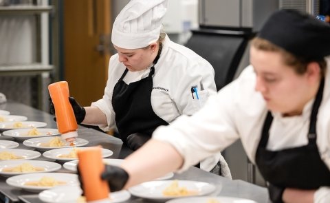 Two Culinary students dressed in chefs' uniforms and hats plate food at an event at Seasoned.