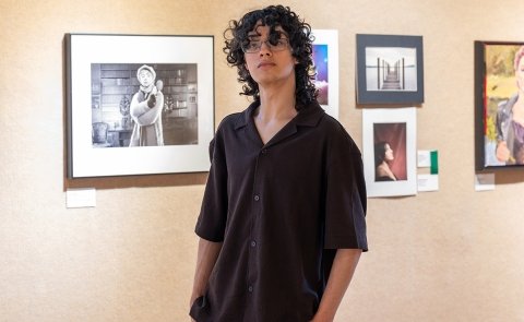 Ivan Aviles stands in front of some of his photography in the 2026 Annual Juried Student Art Exhibition in the Visual Arts Gallery, with his hands in his pockets.